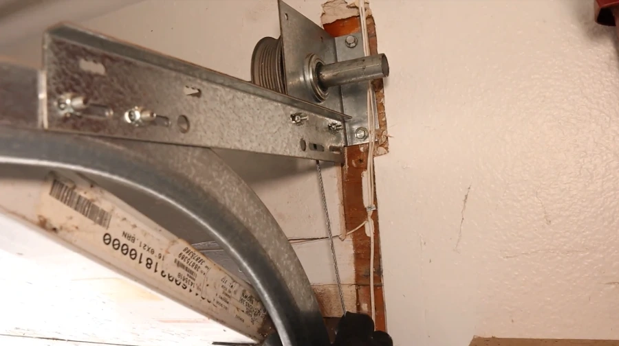 Close-up of a garage door cable drum, end bearing plate and lift cable at the top corner of the door, with the torsion spring label visible