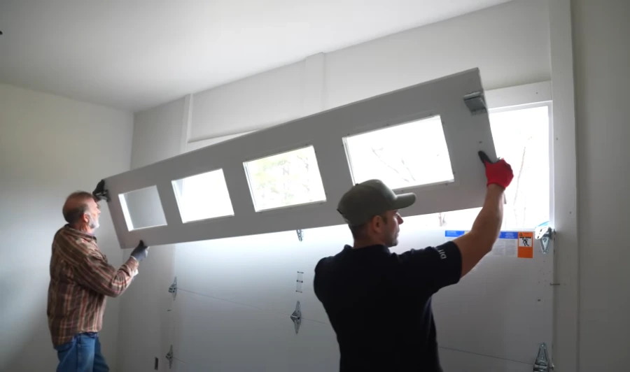 Two technicians lifting a windowed garage door section into place during a panel replacement