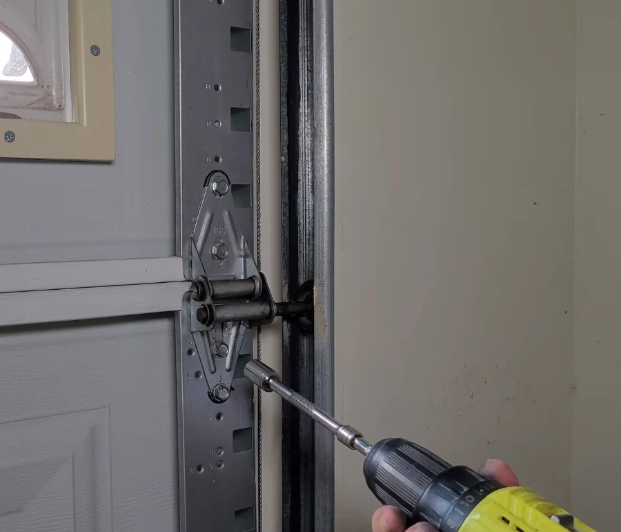 Technician using a cordless drill on a garage door hinge and roller bracket beside the lift cable
