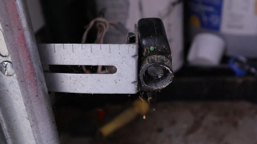 Garage door photo-eye safety sensor mounted low on the track bracket, lens fouled with dust and cobwebs, indicator light showing green